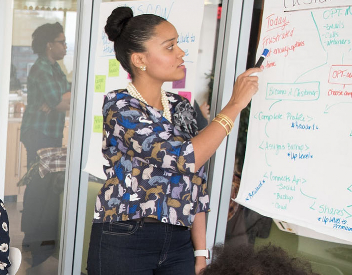 Woman in floral shirt writing on white board while three female collegues work on a table behind her