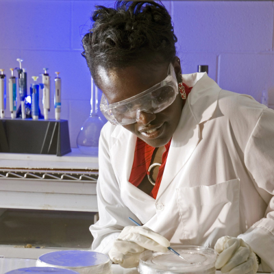 Woman in lab wearing safety goggles examining the contents of a petri dish.