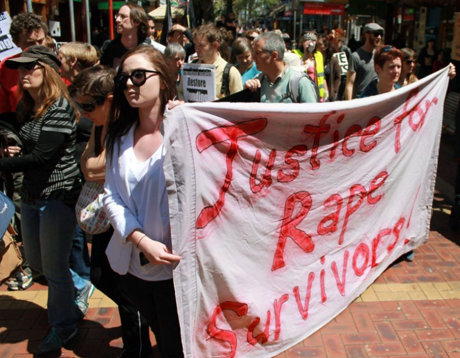 A young woman holds a banner in front of a crowd. The banner reads, Justice for Rape Survivors