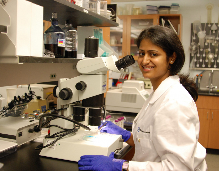 Woman in lab coat and gloves works at microscope in lab.