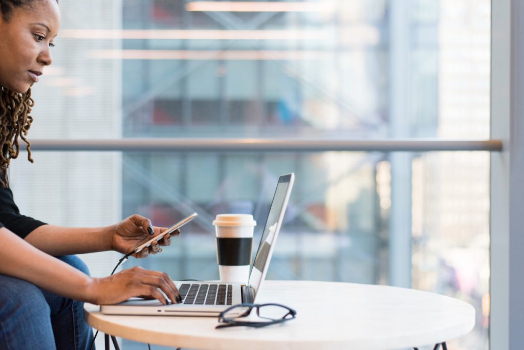 African American women at desk coding on her laptop and using a mobile phone