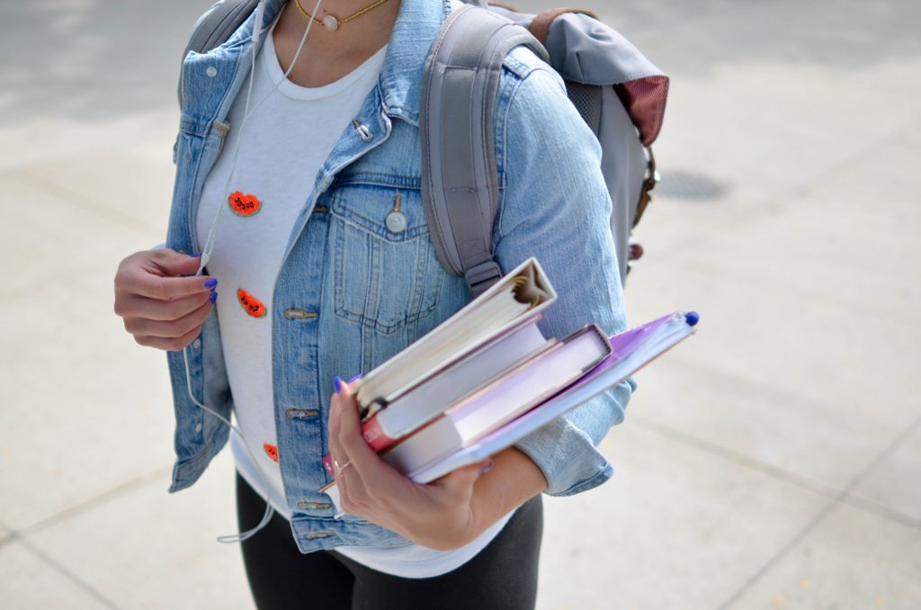 Woman college student in a jean jacket and black pants carrying a backpack, standing and hold an armload of books.