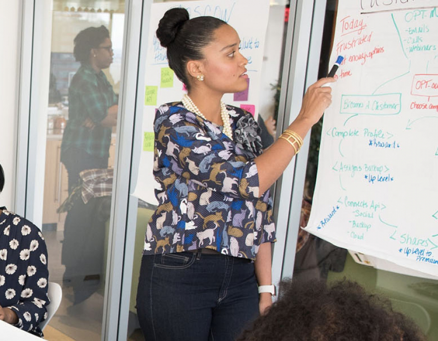 Woman in floral shirt writing on white board while three female collegues work on a table behind her