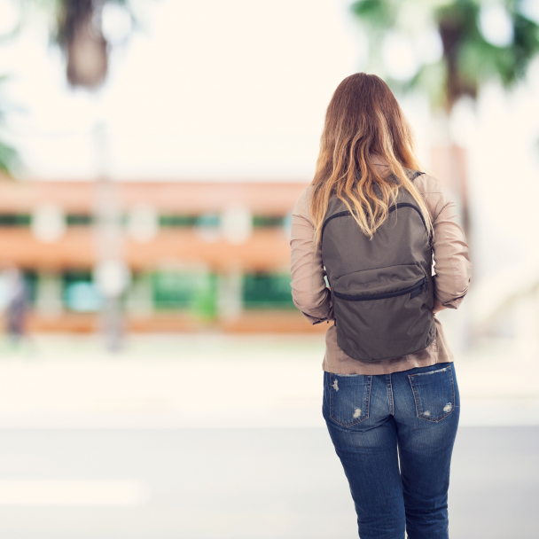 Female student with back to camera walks on college campus with backpack.