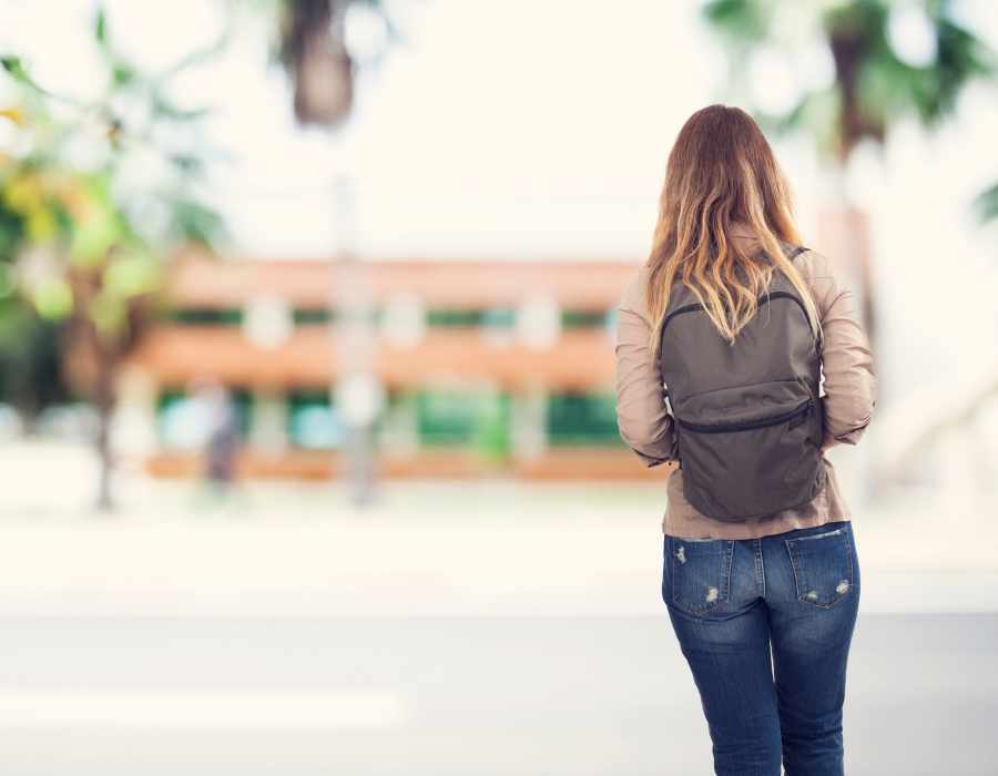 Female student with back to camera walks on college campus with backpack.