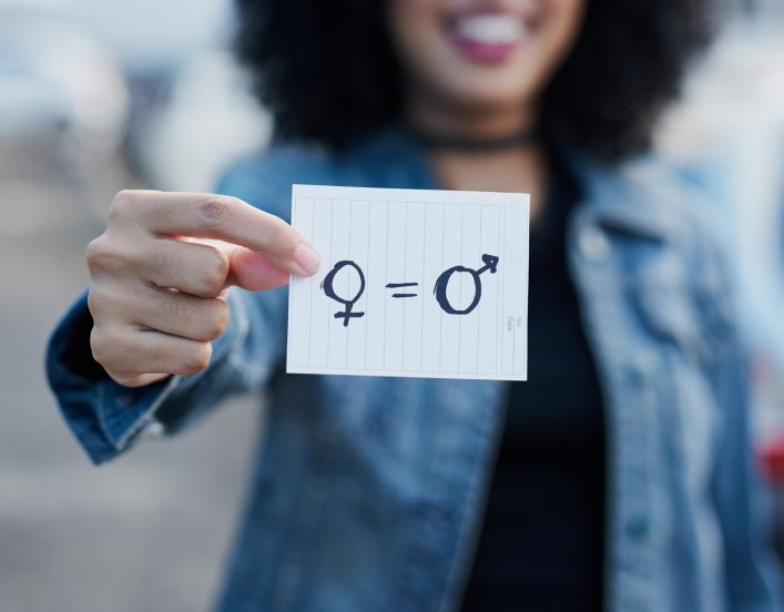 Out of focus woman holding a piece of paper with gender symbols and equals sign drawn on it.