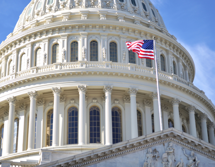 The dome of Capitol Building, Washington DC, and the American flag against a blue sky.