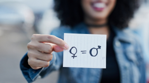 Out of focus woman holding a piece of paper with gender symbols and equals sign drawn on it.