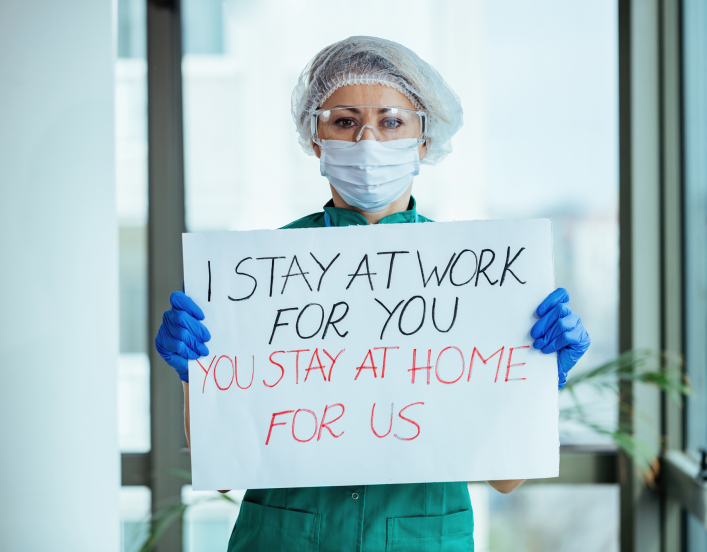 Female doctor holding placard with 'I stay at work for you, you stay at home for us' message during coronavirus pandemic.