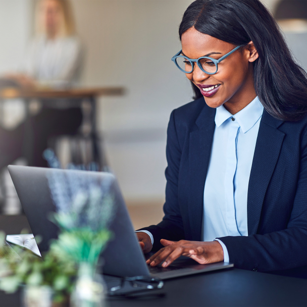 Smiling young Black businesswoman working on a laptop at her desk in a bright modern office with colleagues in the background.