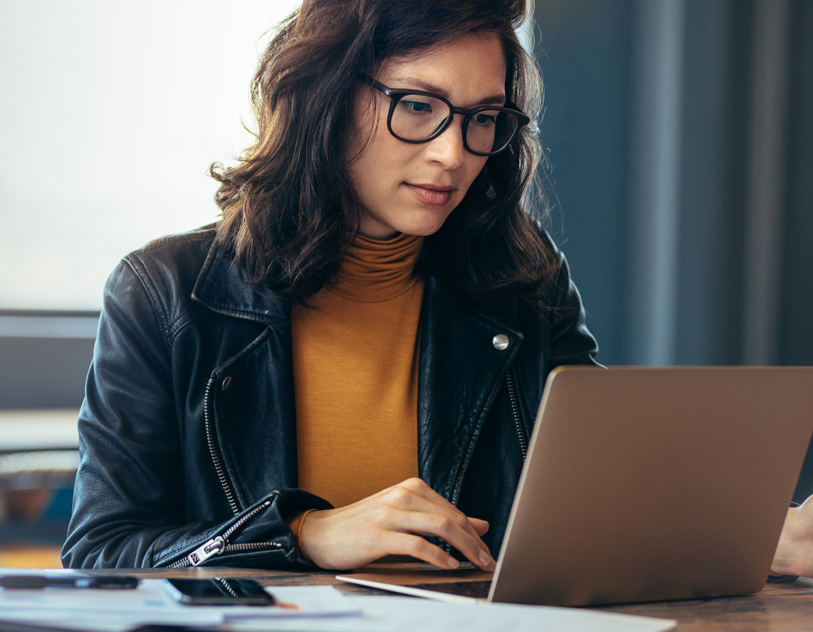 Asian woman wearing glasses and leather jacket types on laptop.