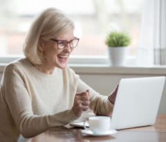 Photo of older woman sitting at table with coffee, smiling at laptop.
