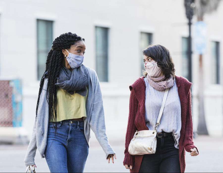 Two multi-ethnic young women in their 20s walking across a city street. They are side by side, looking at each other, conversing. They are wearing protective face masks, trying to prevent the spread of coronavirus during the COVID-19 pandemic.