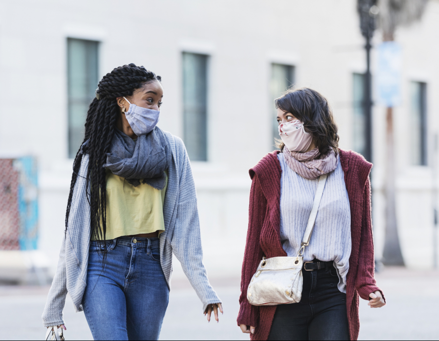 Two multi-ethnic young women in their 20s walking across a city street. They are side by side, looking at each other, conversing. They are wearing protective face masks, trying to prevent the spread of coronavirus during the COVID-19 pandemic.