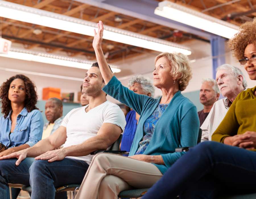 Photo of audience members at a town hall meeting. Woman raises hand.